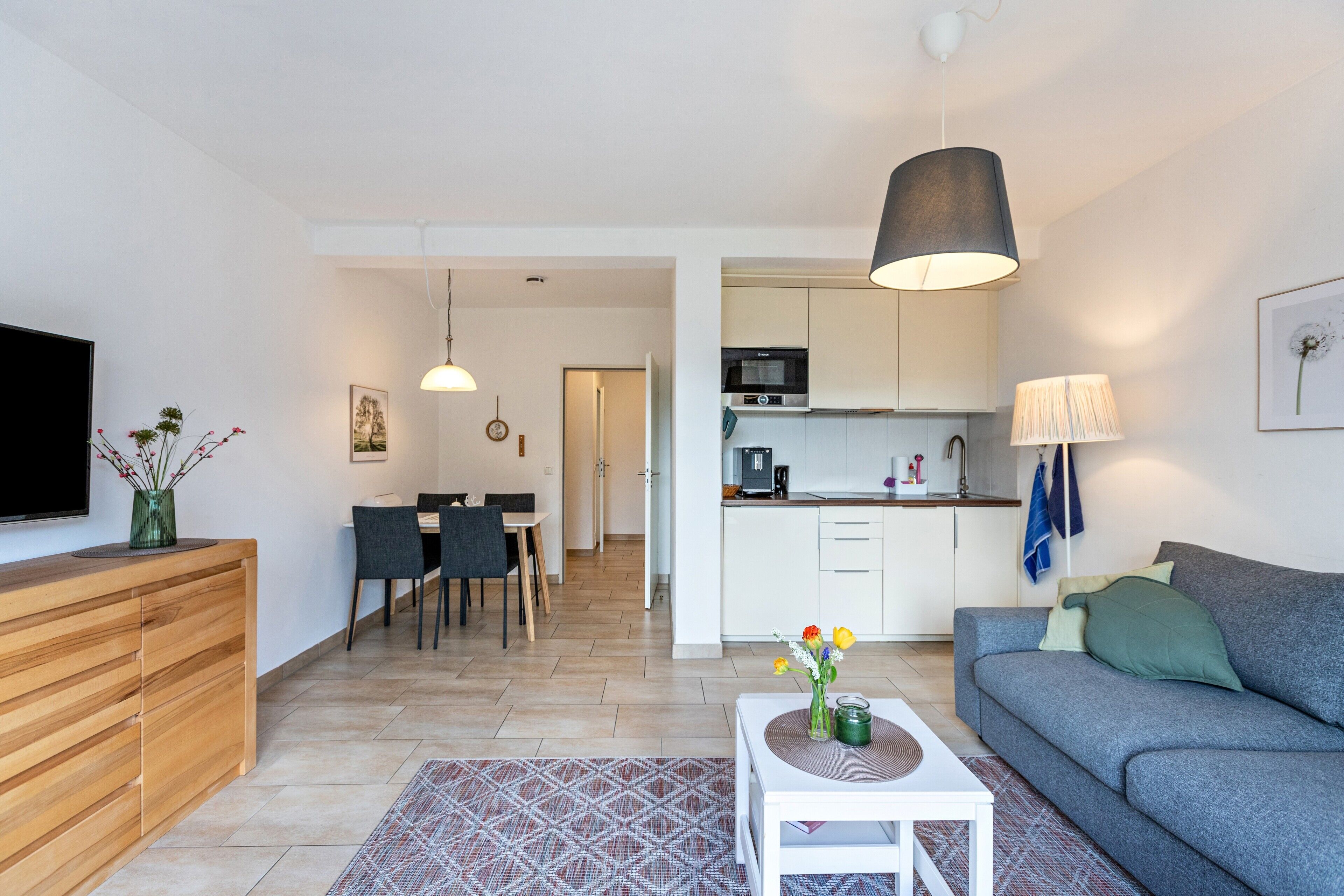Ein modernes Wohnzimmer mit einer Couch, einem Fernseher, einem Holzschrank, einem Essbereich mit Stühlen und einer Küche mit Schränken und Geräten.