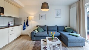 Un salon moderne avec un canapé gris, une table basse blanche et un coin cuisine doté de meubles blancs et d’une lampe suspendue.