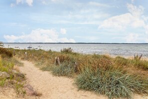 Uma trilha de areia que leva até a água, com barcos ao longe.