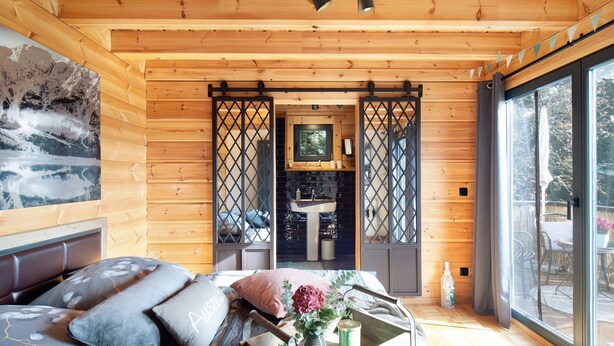 L’intérieur d’une cabane en bois, avec un lit, une fenêtre, un évier et une porte donnant sur l’extérieur.