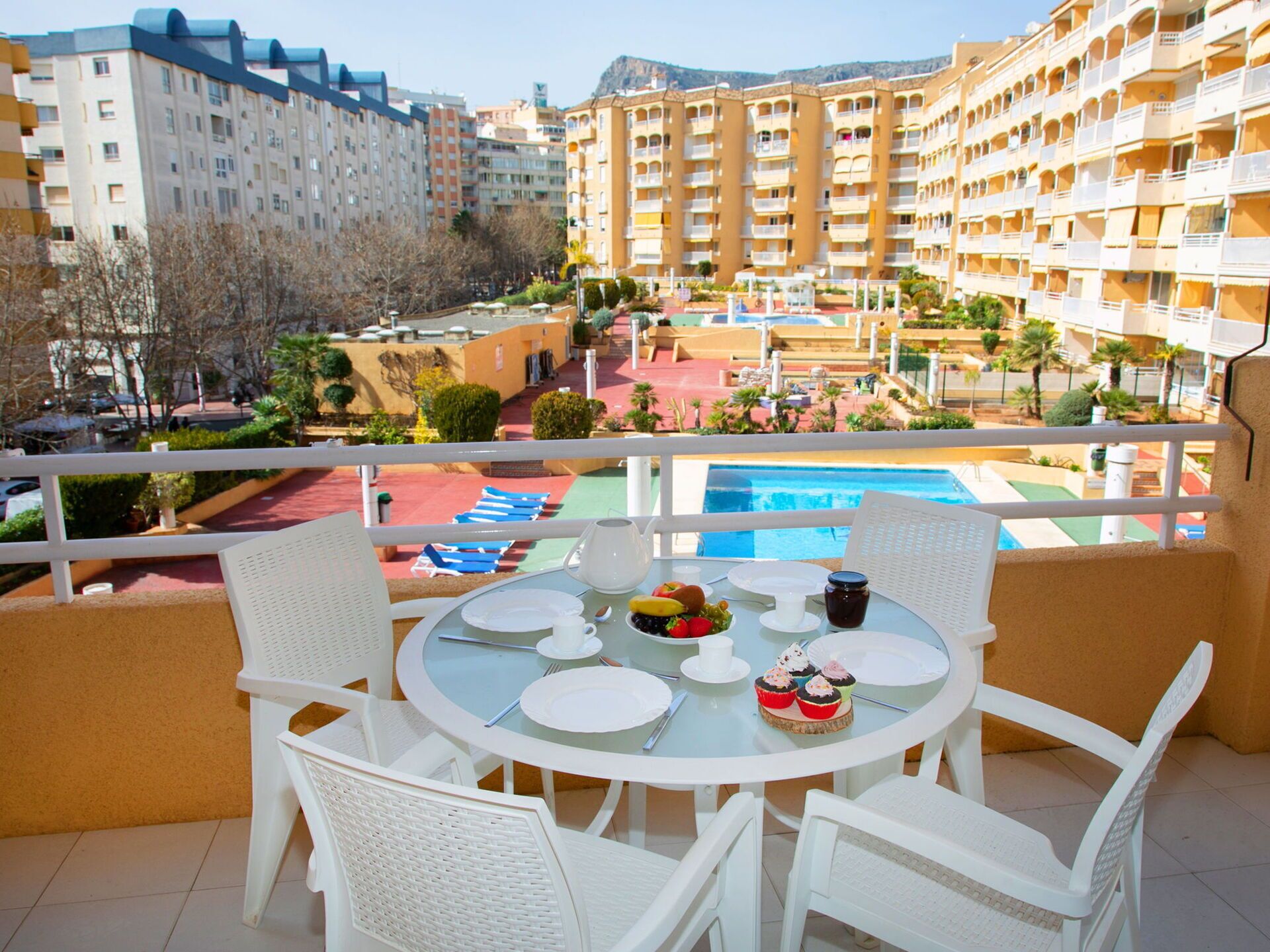 A balcony with a round table set for dining, overlooking a pool and surrounding buildings.