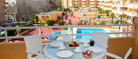 A balcony with a round table set for dining, overlooking a pool and surrounding buildings.