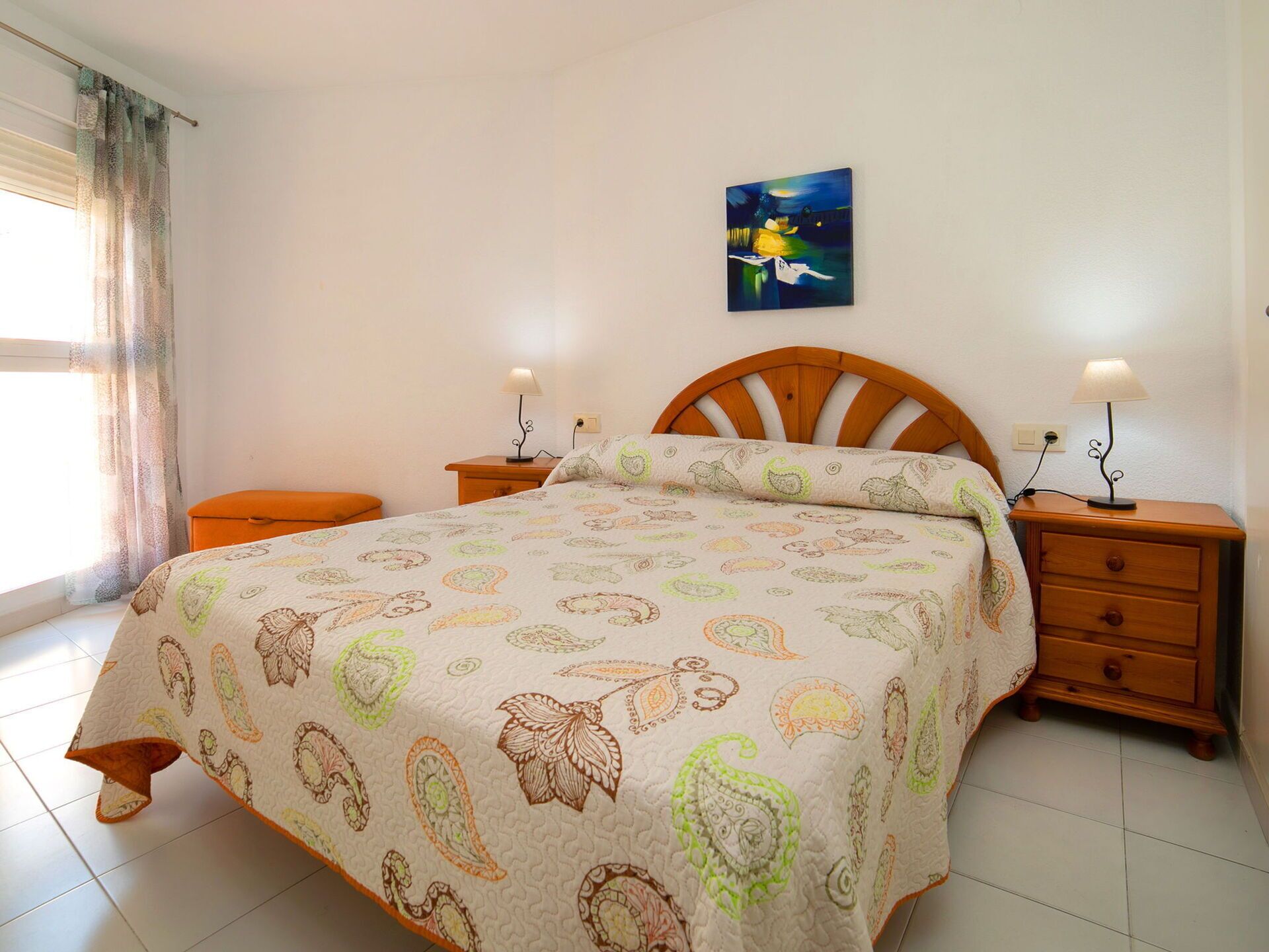 A bedroom with a bed, two bedside tables, a nightstand, a painting on the wall, and a window with curtains.