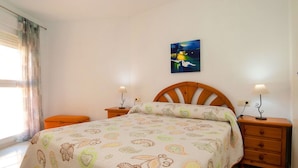 Ein Schlafzimmer mit einem Bett, zwei Nachttischen, einem Schranken und einem Bild an der Wand, sowie einem Fenster mit Vorhängen.