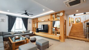A modern living room with a sofa, ottomans, a glass coffee table, a TV, wooden shelves, and a staircase.