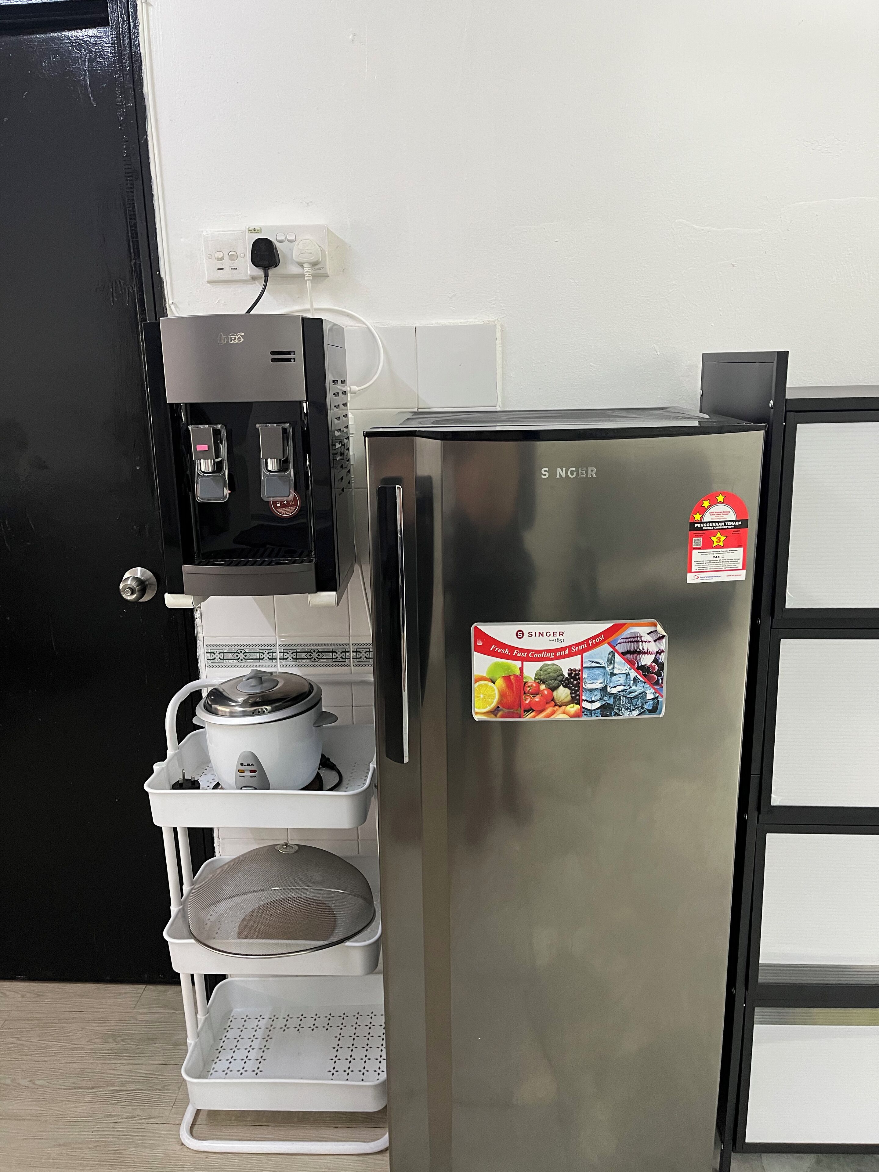 A stainless steel refrigerator with a sticker, a coffee machine, and a white shelf unit with a pot and strainer.