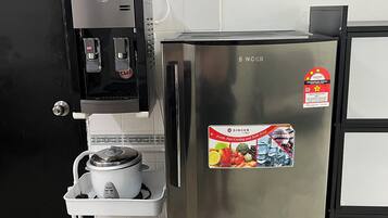 A stainless steel refrigerator with a sticker, a coffee machine, and a white shelf unit with a pot and strainer.