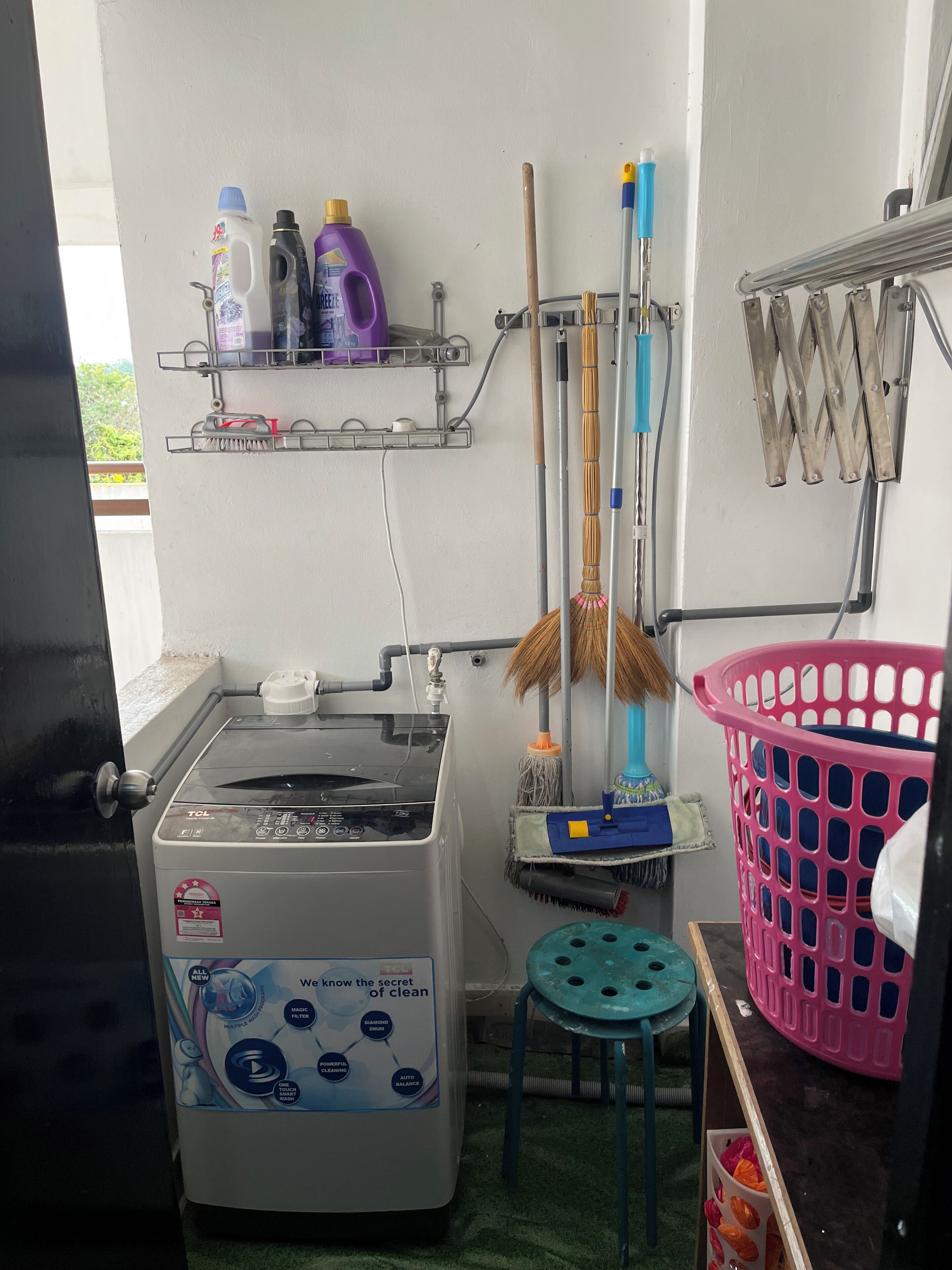 A laundry area with a washing machine, shelves with cleaning supplies, a mop, a broom, and a pink laundry basket.
