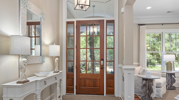 Uma entrada ampla com porta de madeira, um console branco com abajur e vista para o verde lá fora.