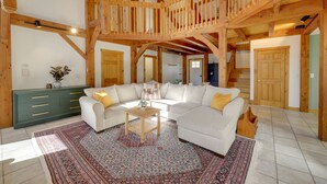 Ein geräumiges Wohnzimmer mit hoher Holzbalkendecke, einem großen, weißen Modulsofa, einem grünen Schrank und einem gemusterten Teppich.