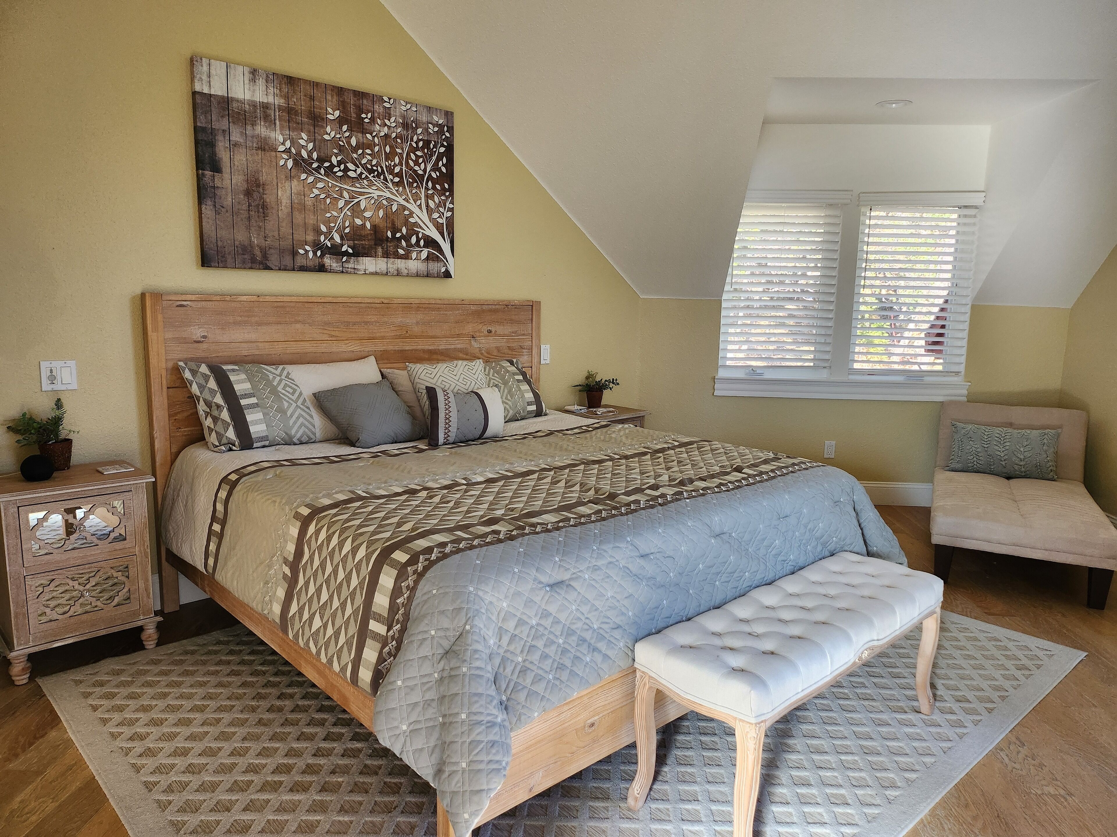 A bedroom with a wooden bed, a bench, a nightstand, a potted plant, and a window with blinds.