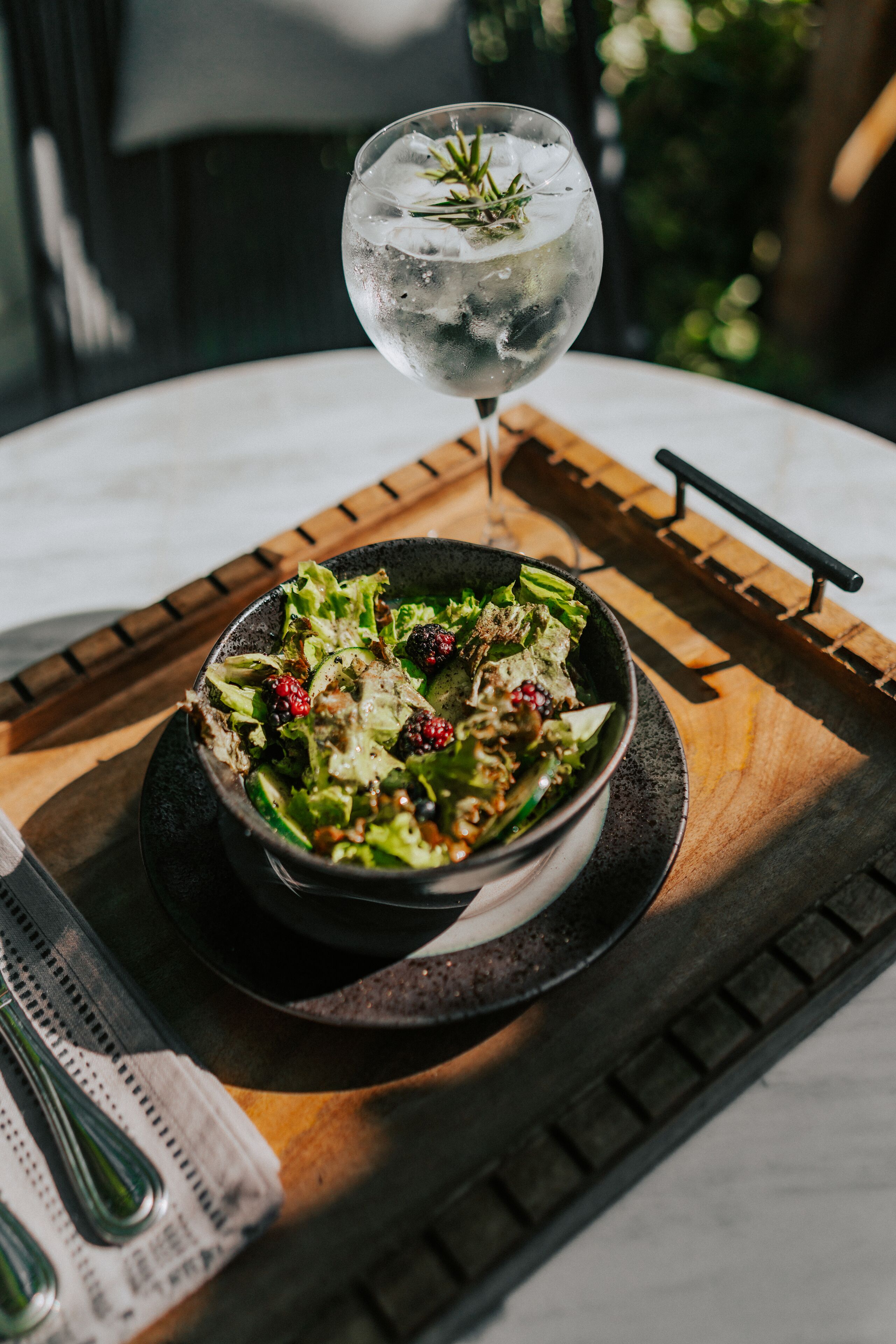 A bowl of salad with blackberries and a glass of iced water with a sprig of rosemary on a wooden tray.
