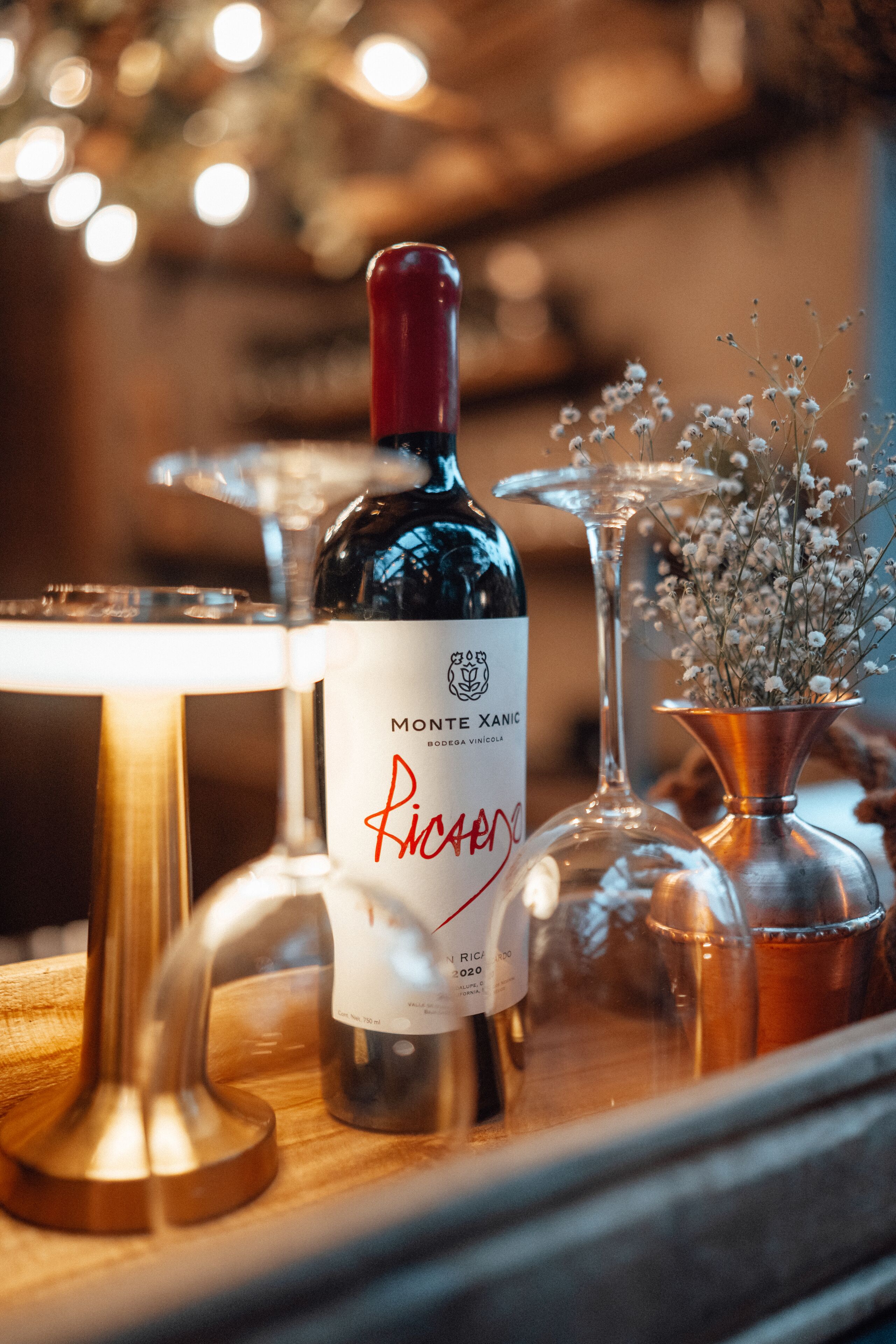 A bottle of Monte Xanic wine with a label, placed on a wooden surface with a glass and a copper vase with flowers.