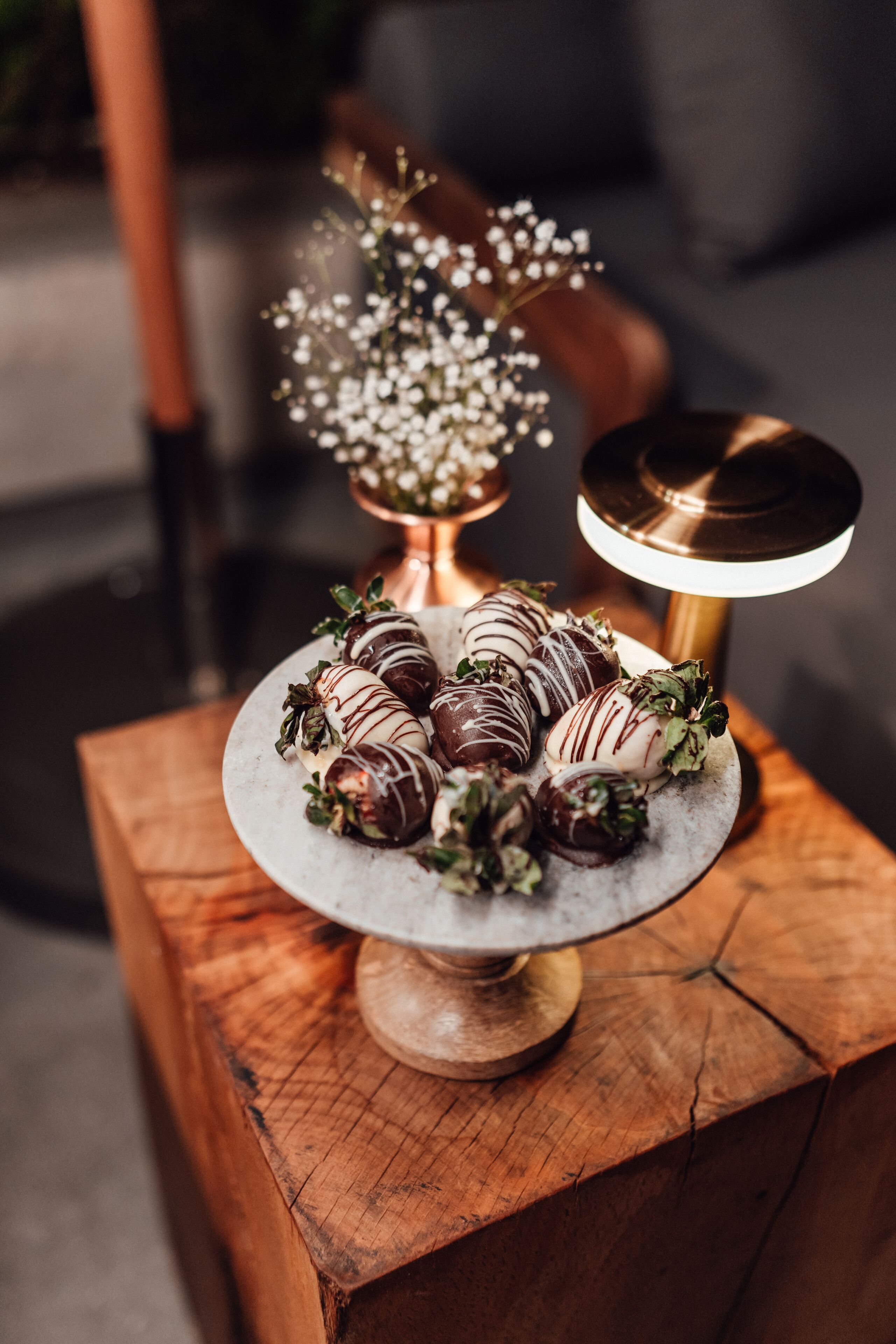 A plate of chocolate-covered strawberries on a marble stand with a small vase of white flowers in the background.