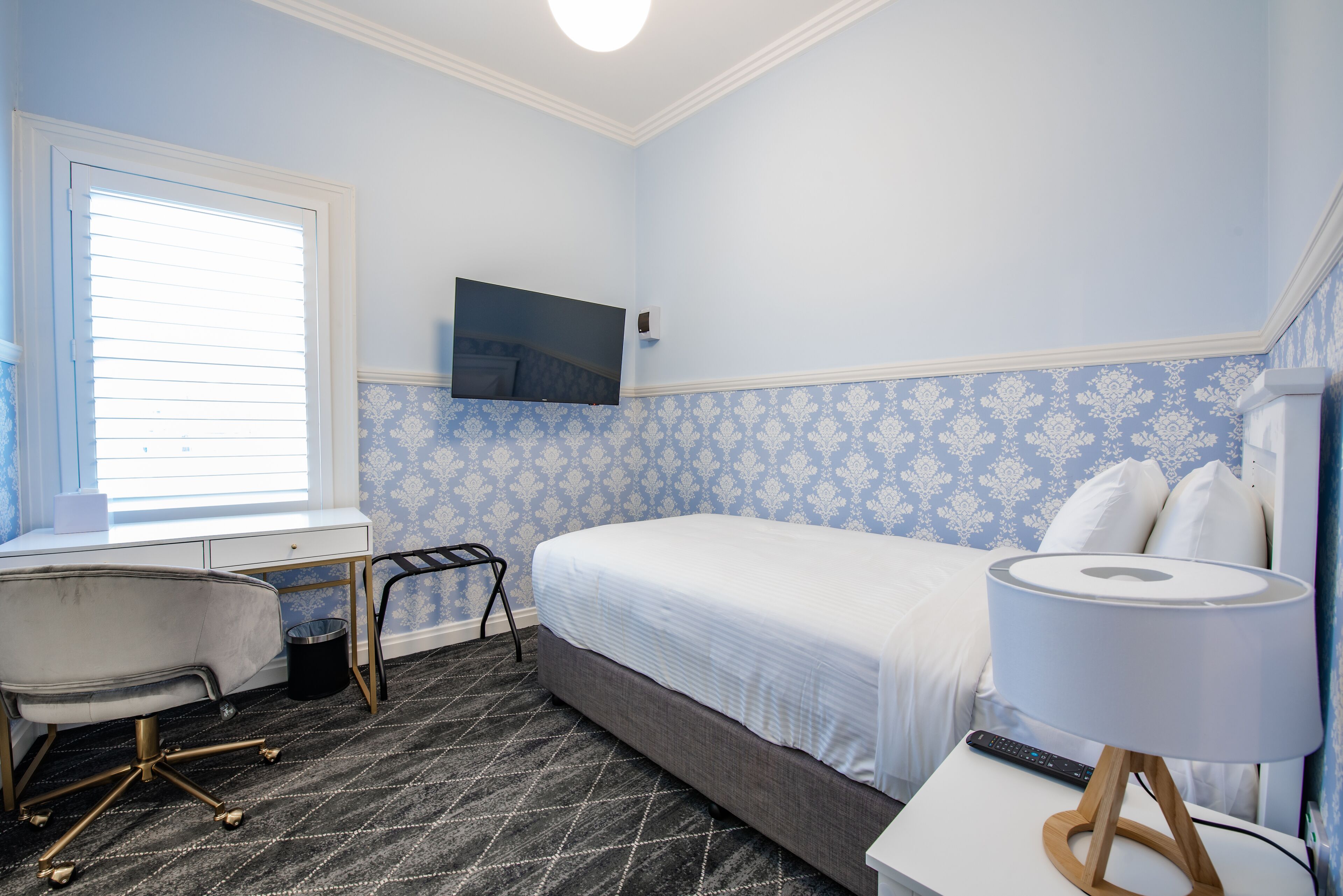 Ett hotellrum med en säng, ett skrivbord med en stol, en tv som är monterad på väggen och en lampa på ett nattduksbord.