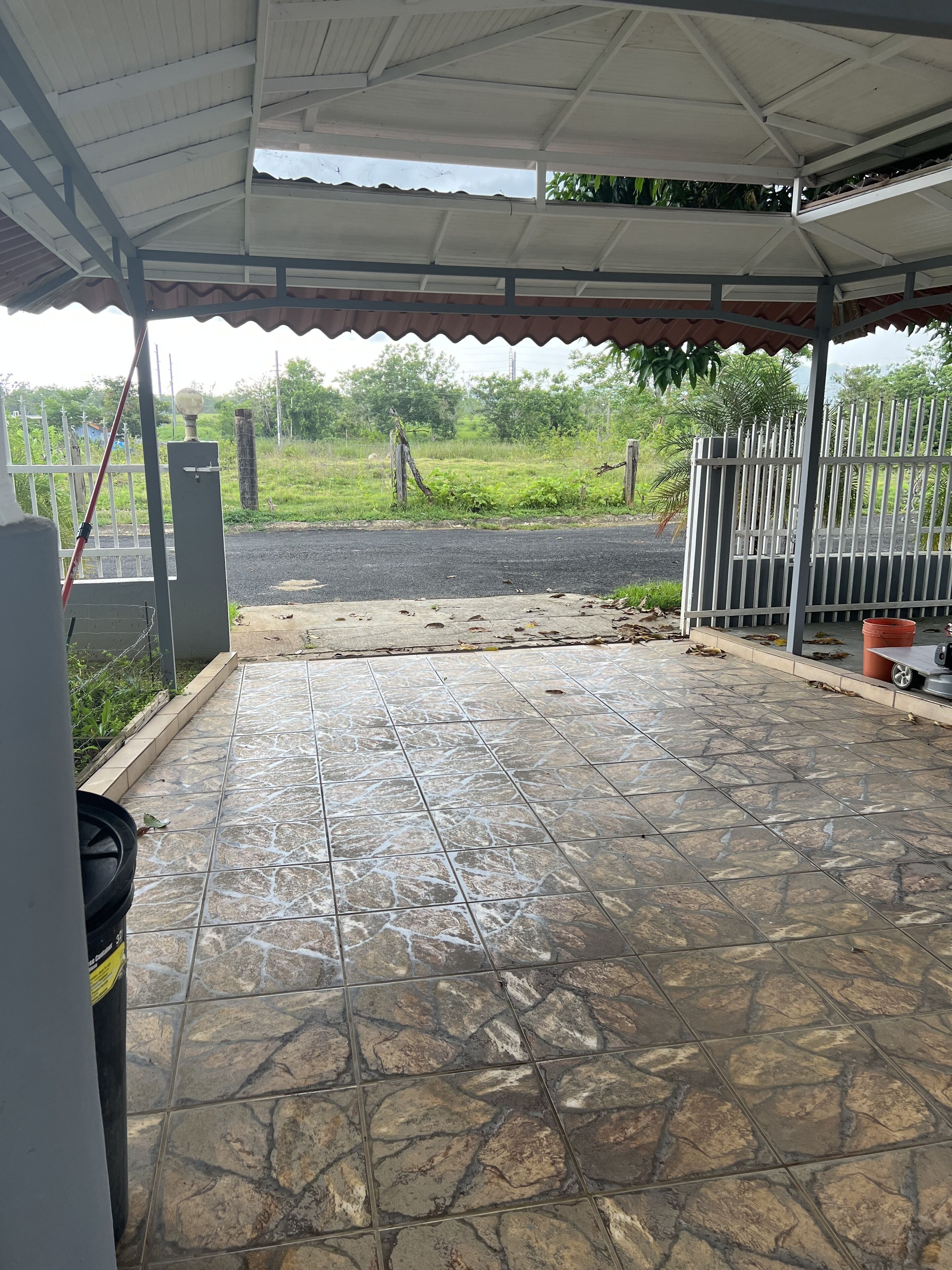 A covered outdoor area with a tiled floor, a metal gate, and a view of a road and greenery.