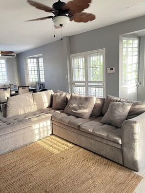 Una sala de estar con sofá modular, ventilador de techo y ventanas amplias.