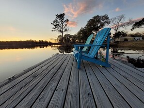 Eine Holzsteg mit einem blauen Adirondack-Stuhl, der auf einen stillen See im Sonnenuntergang blickt.