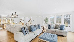 Un salón amplio con un conjunto de sofás, un reposapiés y una zona de comedor con una cocina al fondo.