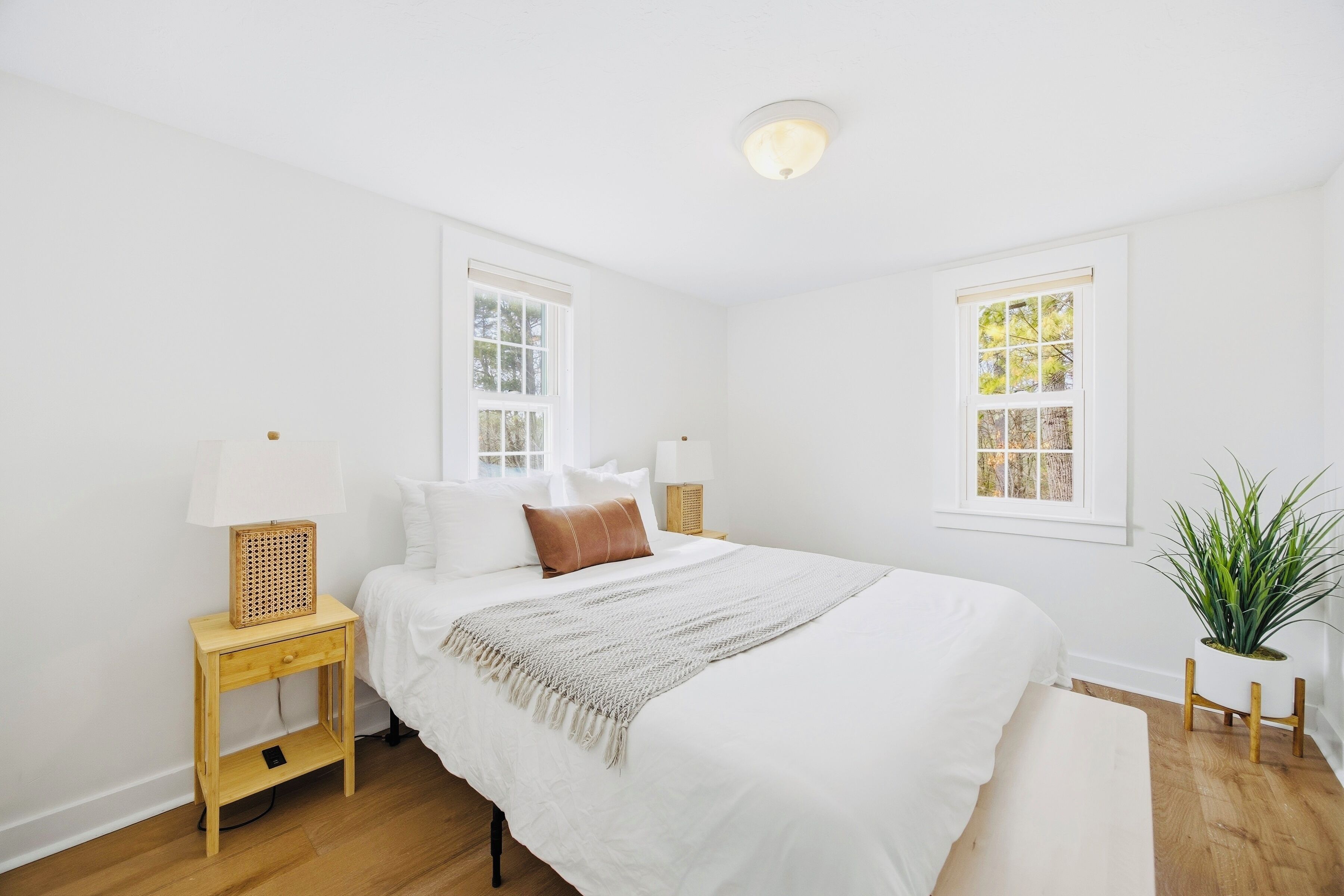 A bedroom with a bed, two bedside tables, a lamp, a bedside plant, and a window.