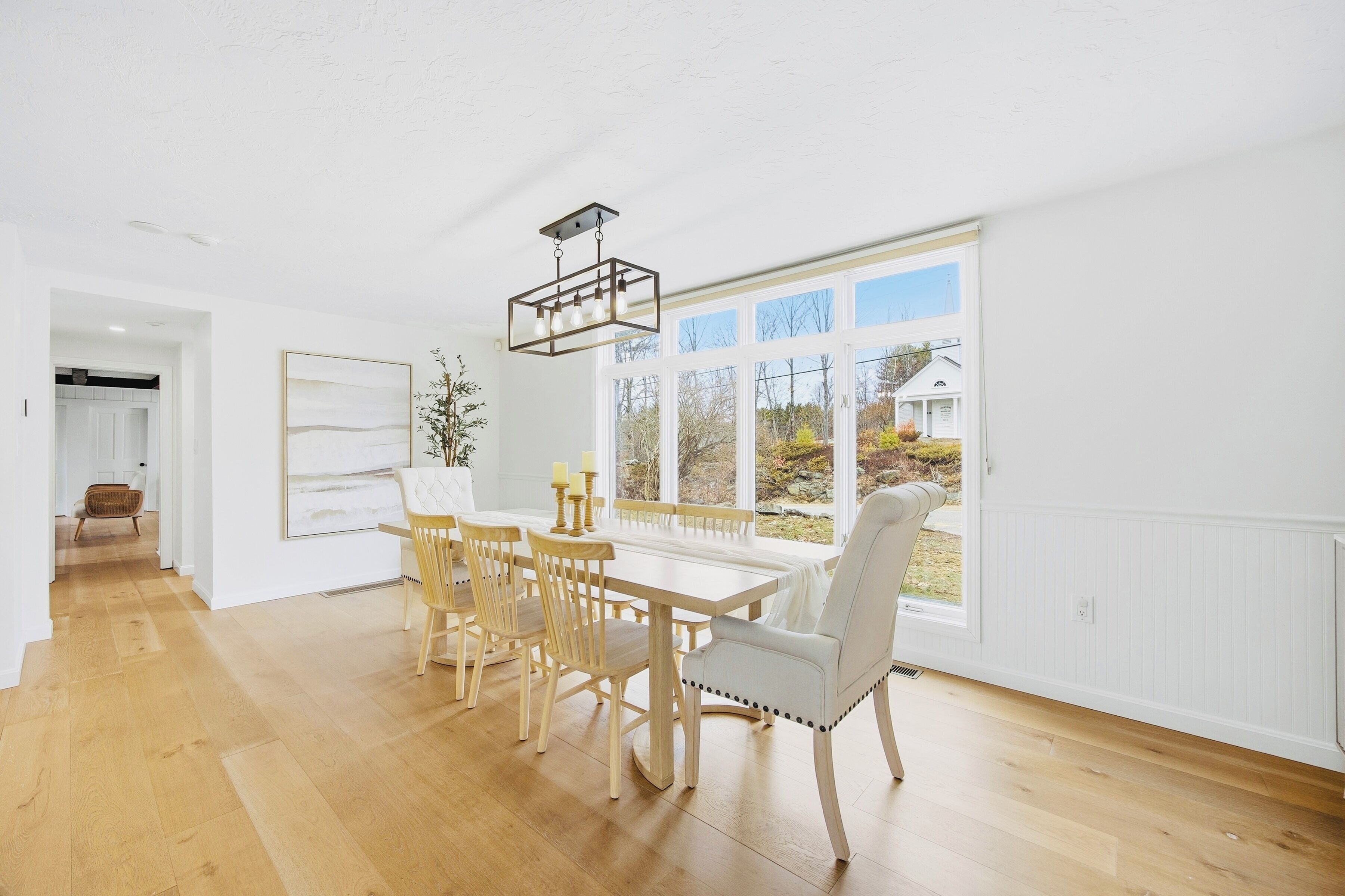 Un espace repas avec une table et des chaises en bois, un luminaire suspendu et une grande fenêtre donnant sur l’extérieur.