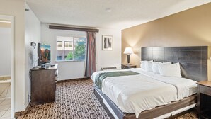 A hotel room with a bed, a desk, a television, and a window with curtains. - Coratel Suites Deluxe 1 King Suite with Sofa Oversized Non Smoking (Wichita)