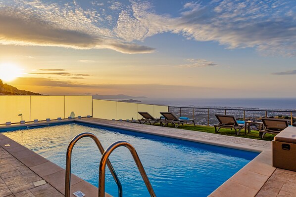 A swimming pool with lounge chairs and a view of the ocean at sunset.
