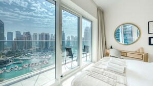 A modern hotel room with a large balcony, a bed with white linens, a round mirror, and a view of a marina and city skyline. - Maison Privee - Aesthetic Apt w/ Stunning Vw in Luxurious Building (Dubai)