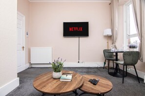 A room with a TV displaying 'NETFLIX,' a potted plant, a book, a remote control, and a dining area with chairs and a table.
