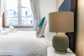 A neatly made bed with rolled towels, a textured green lamp on a bedside table, and a window with curtains.