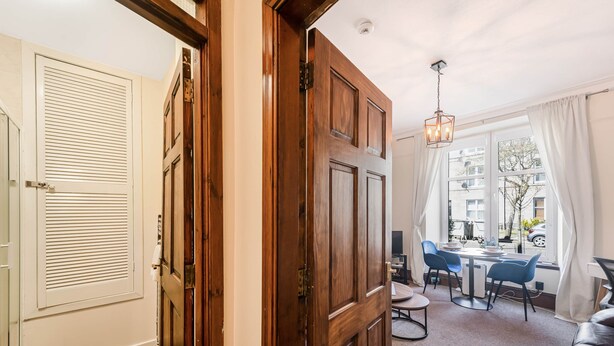 Una habitación con puerta de madera, ventana con cortinas, mesa redonda con sillas y sofá de cuero.