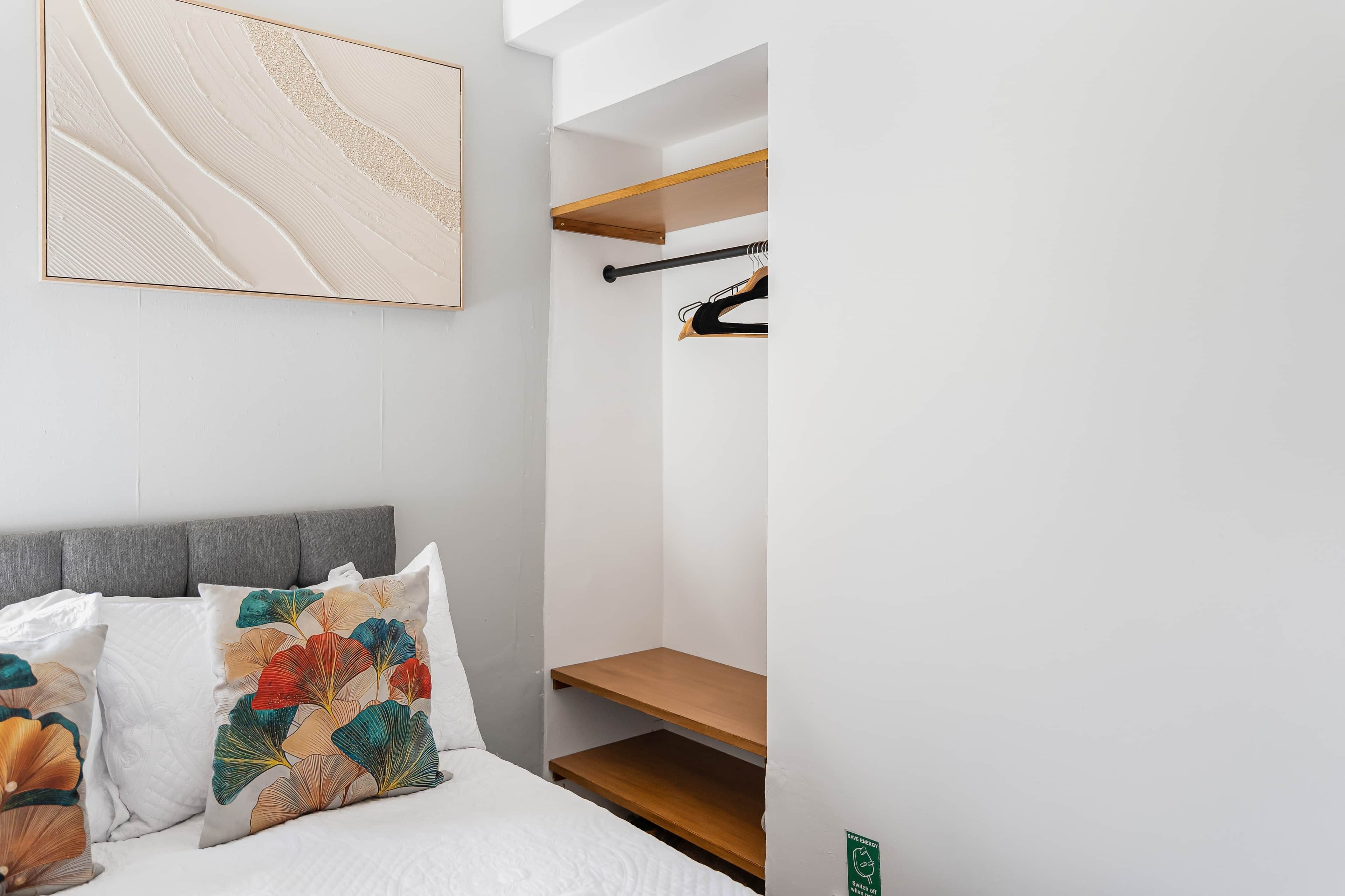 A bedroom with a bed, pillows, a wall-mounted artwork, a coat rack, and a wooden shelf.
