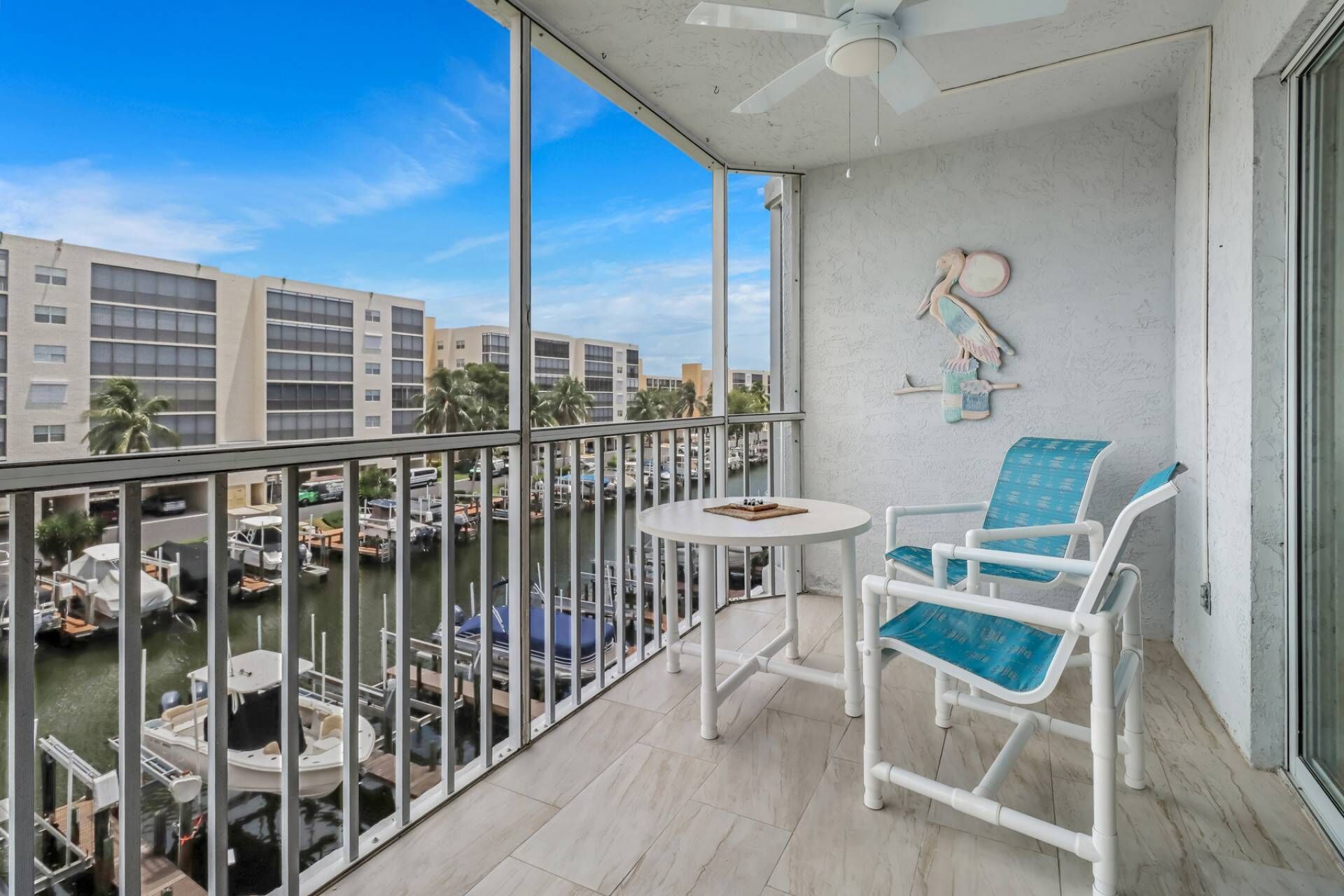 A balcony with a view of a marina, white chairs, a small table, and a ceiling fan.