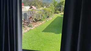 A view through a window with curtains, featuring a well-maintained lawn, a small garden with various plants, and a distant view of a house and trees. - Modern 2 bedroom Unit in the beautiful Scenic Rim (Rathdowney)