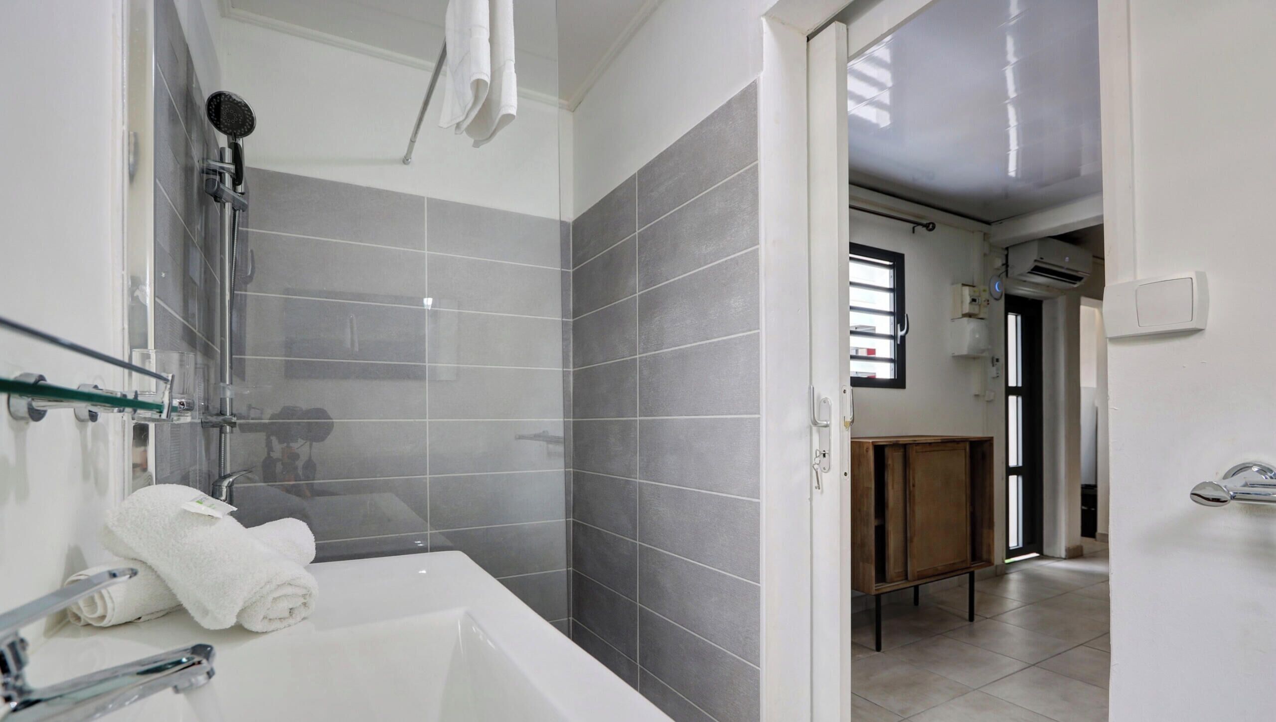 Un baño moderno con bañera, ducha y baldosas grises. Hay un armario de madera y una repisa de vidrio.