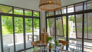 A dining area with a round glass table set for four, surrounded by large windows with blinds, a woven light fixture, and a view of greenery outside.
