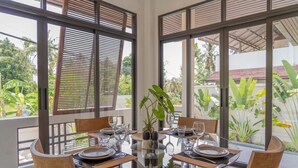A round glass table set for four with wooden chairs, surrounded by large windows with wooden blinds, overlooking a tropical garden.