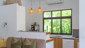 A modern kitchen with a dishwasher, a window with a view of greenery, a wooden countertop, and a beige sofa with cushions.