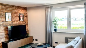 A modern living room with a brick wall, a flat-screen TV, a round coffee table, a sofa, and a window with curtains. - Industrial Loft Urban. Living. Trier (Trier)