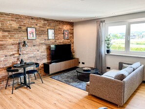 A living room with a brick wall, a flat-screen TV, a sofa, and a dining area with chairs and a table. - JUPAS° Loft34 – Studio, Carport & Workspace (Trier)