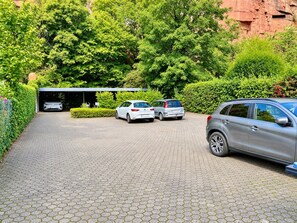Ein gepflasterter Weg führt zu einem überdachten Parkbereich mit drei Autos, umgeben von üppiger Vegetation und einer roten Felsformation im Hintergrund.