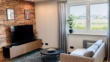 A living room with a brick wall, a flat-screen TV, a sofa, a coffee table, and a window with curtains.
