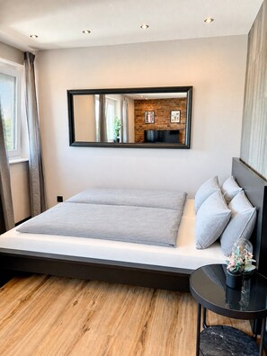 A modern bedroom with a large bed, a small side table, and a mirror on the wall. - JUPAS° Loft34 – Studio, Carport & Workspace (Trier)