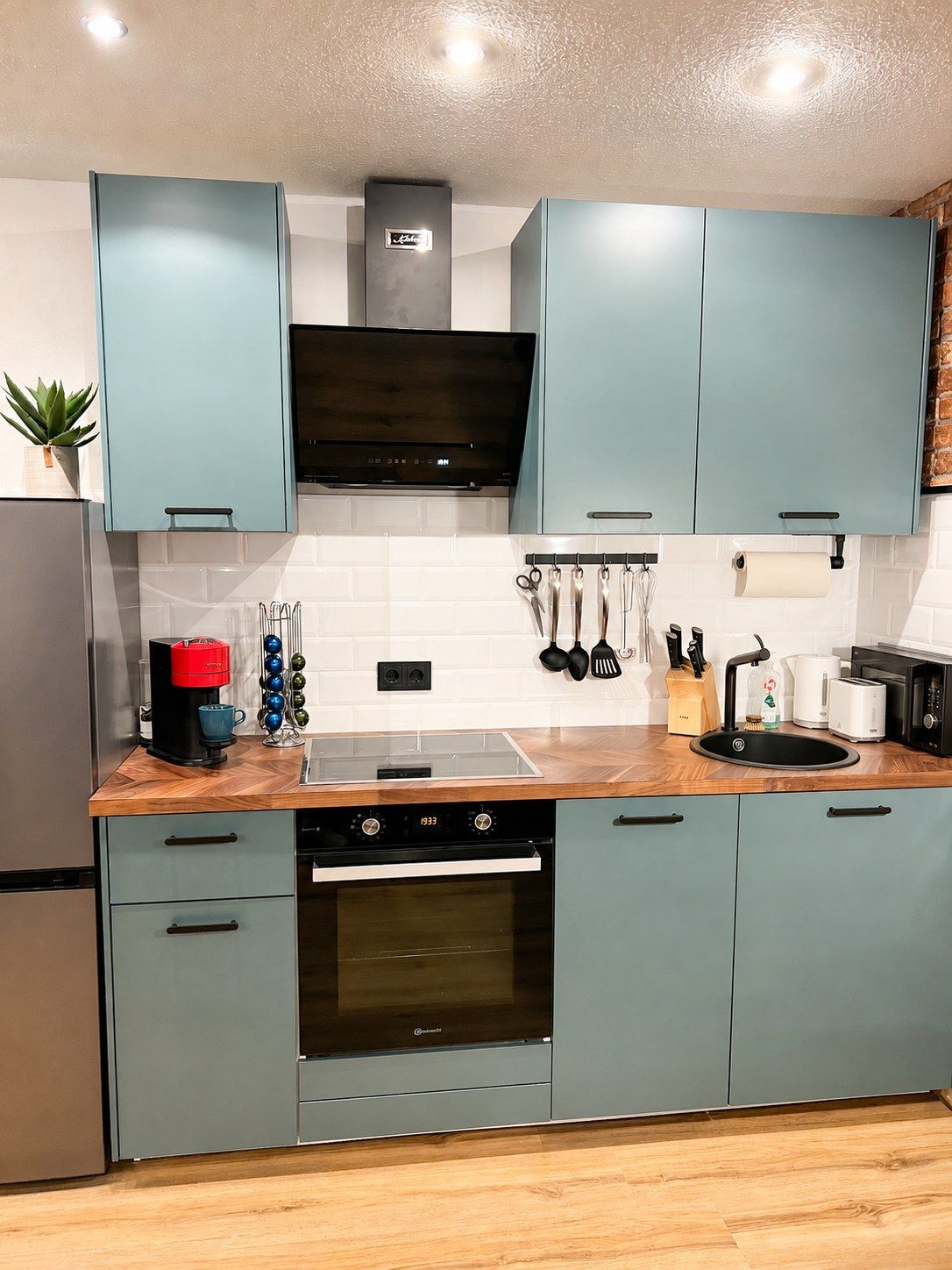 A modern kitchen with light blue cabinets, a black range hood, and a stainless steel refrigerator.