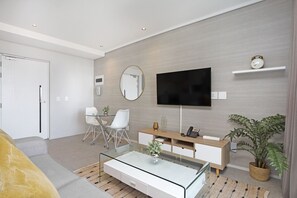 A modern living room with a glass coffee table, a sofa, a TV, a desk, and a plant.
