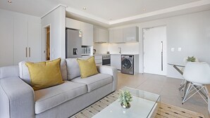 A modern living room with a grey sofa, a glass coffee table, and a kitchen in the background. - Cosmopolitan 401 (Cape Town)