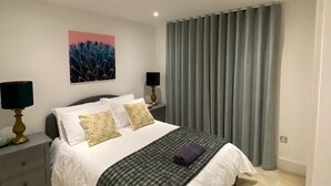 A modern bedroom with a large bed, two bedside tables with lamps, a wall-mounted painting, and a window with grey curtains.