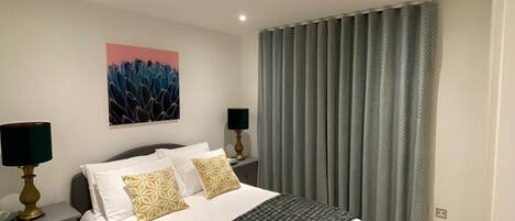 A modern bedroom with a large bed, two bedside tables with lamps, a wall-mounted painting, and a window with grey curtains.