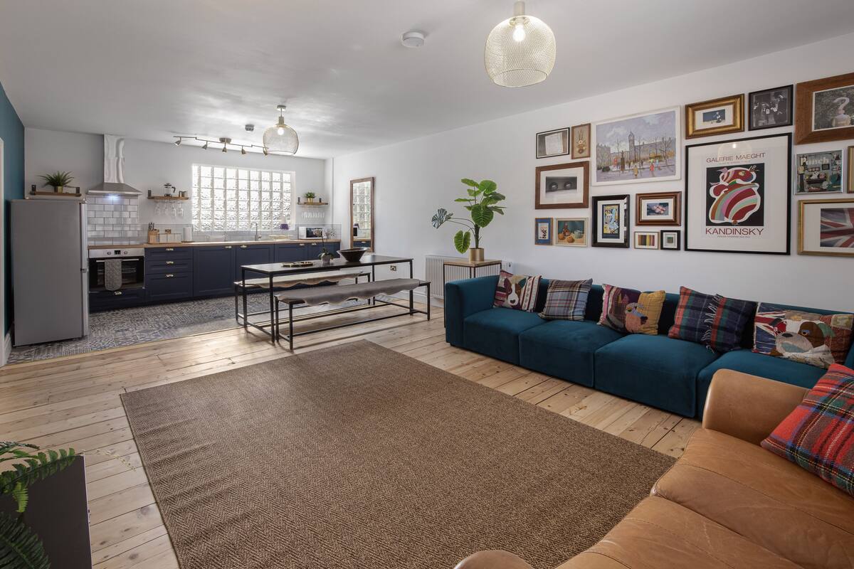 A modern living room with a sofa, coffee table, and a rug. There is a kitchen in the background with a window, and the walls are decorated with framed pictures.