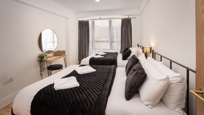 A hotel room with two beds, a bedside table with a lamp, a mirror, and a window with curtains. - Blueberries Apartments by Sasco (Blackpool)
